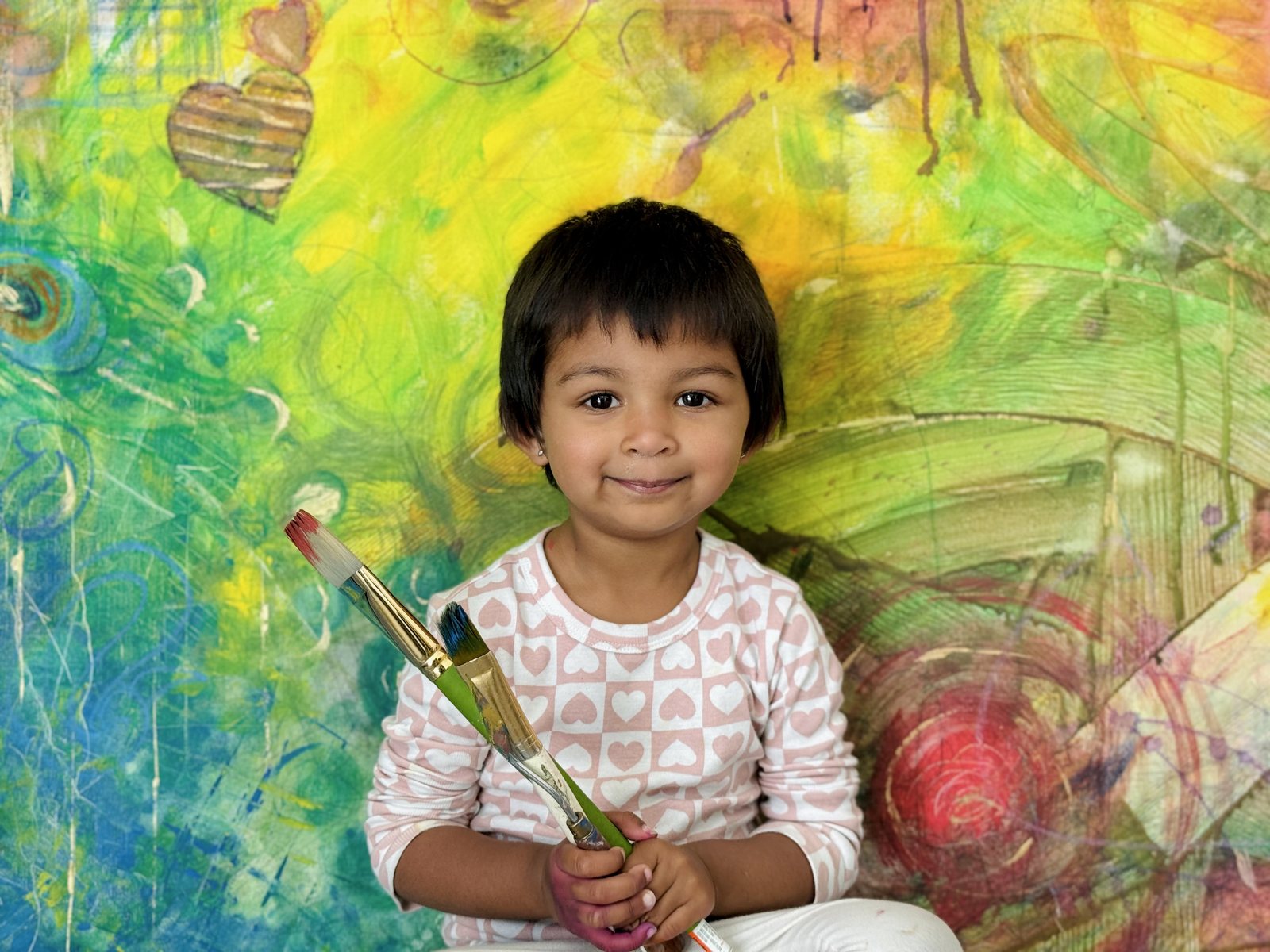Stavya sitting in front of her large colorful painting, holding two paintbrushes